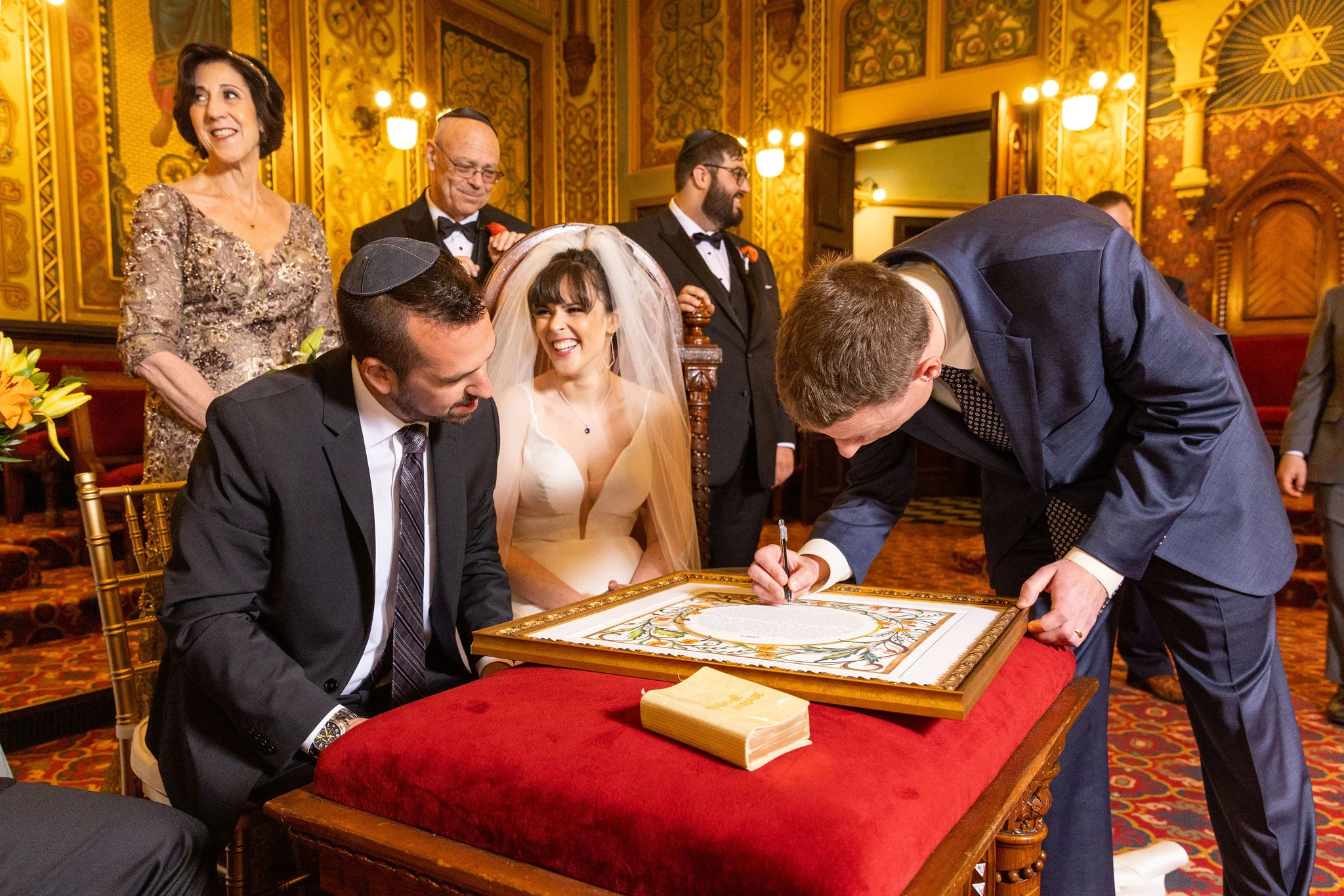 Bride and groom seated while man signs framed document