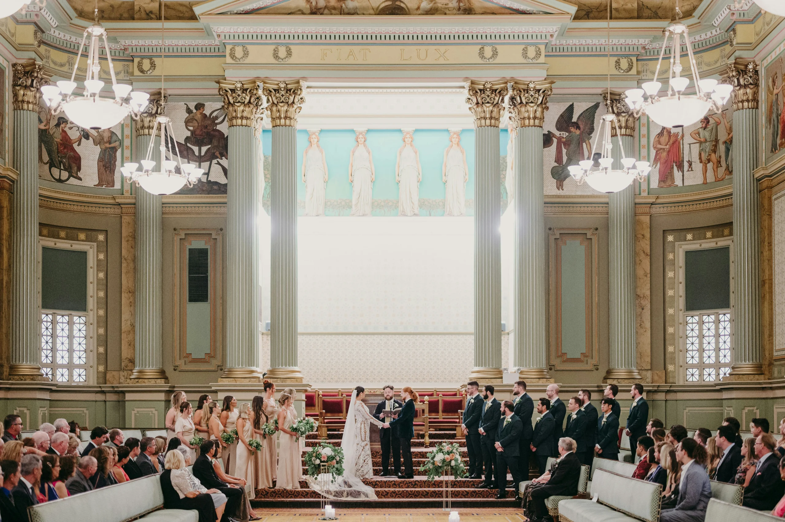 Couple getting married in brightly decorated room surrounded by wedding party and guests