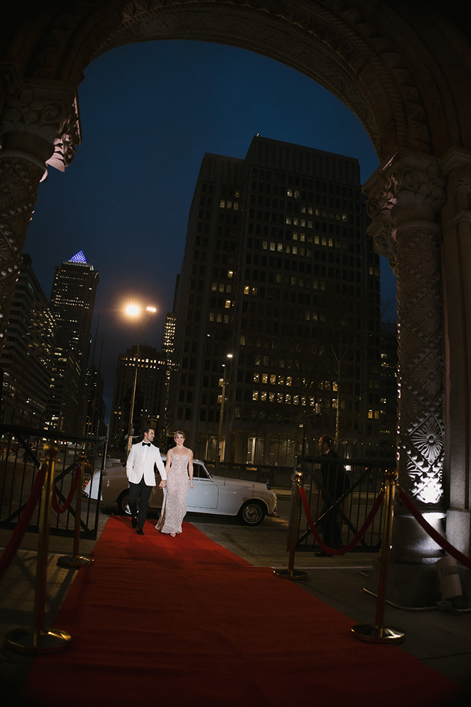 12_31_24_J_M_Wedding_-697sm Bride and groom walking down red carpet together framed by building archway at night.