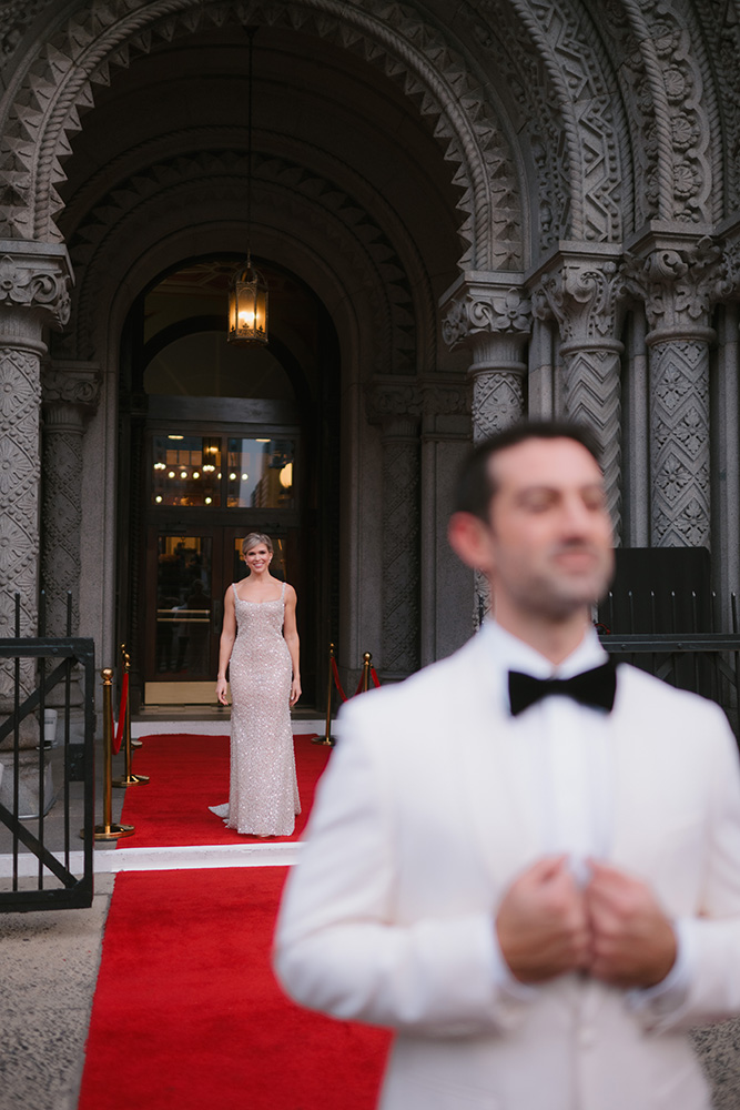 12_31_24_J_M_Wedding_-776sm Smiling bride at the end of a red carpet with groom standing in the foreground, looking away.