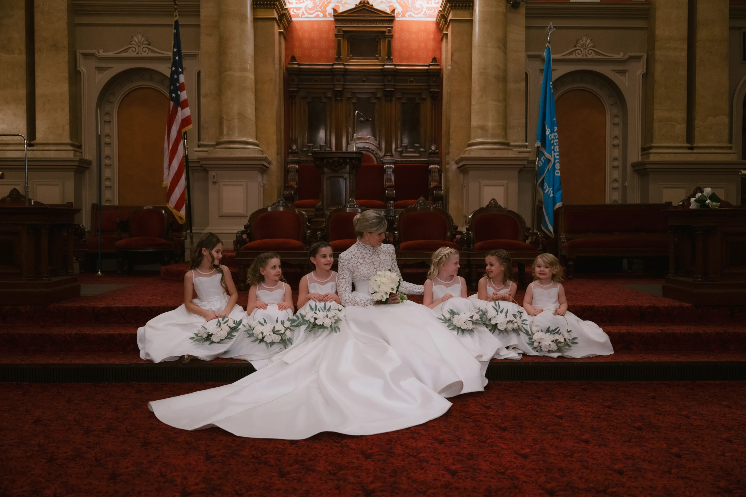 12_31_24_J_M_Wedding_-1054 Bride seated on red, carpeted stairs surrounded by young girls with white dresses and flowers.