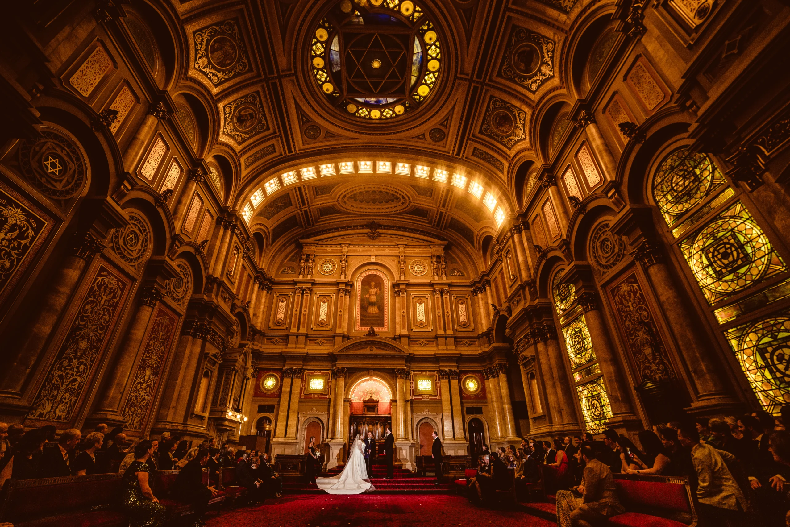 182122DSC09251 Sprawling room with column-decorated walls and stained glass windows. A bride and groom are standing at the front of the room and guests are seated on either side.