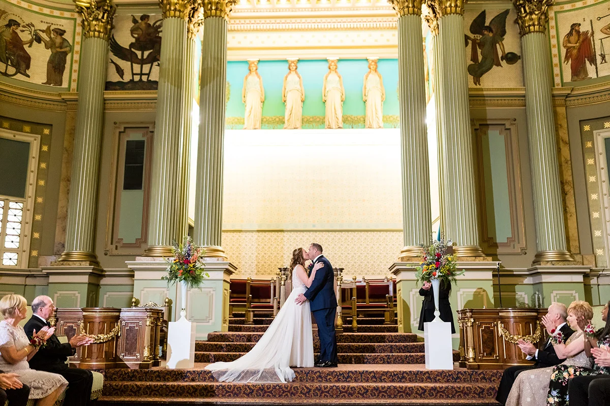 190810BI-372 Bride and groom kissing on staircase while guests clap. Behind them are Grecian-inspired murals and sculptures on the walls.