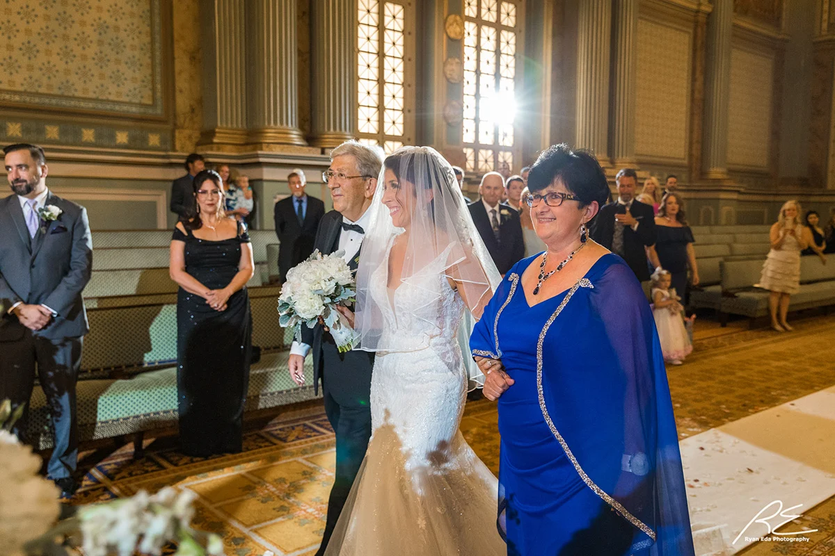 2021-07-24-W-MacIntosh-324 Smiling bride walking up the aisle with her parents. Guests standing in a large, ornate room can be seen behind her.