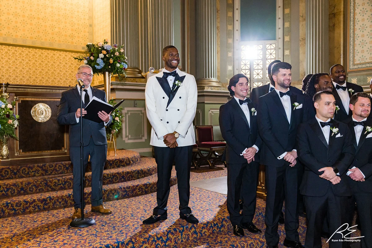 2022-10-01-Akintoye-Wedding-365 Smiling groom, officiant and groomsmen standing together while window and columns are behind them in the background.