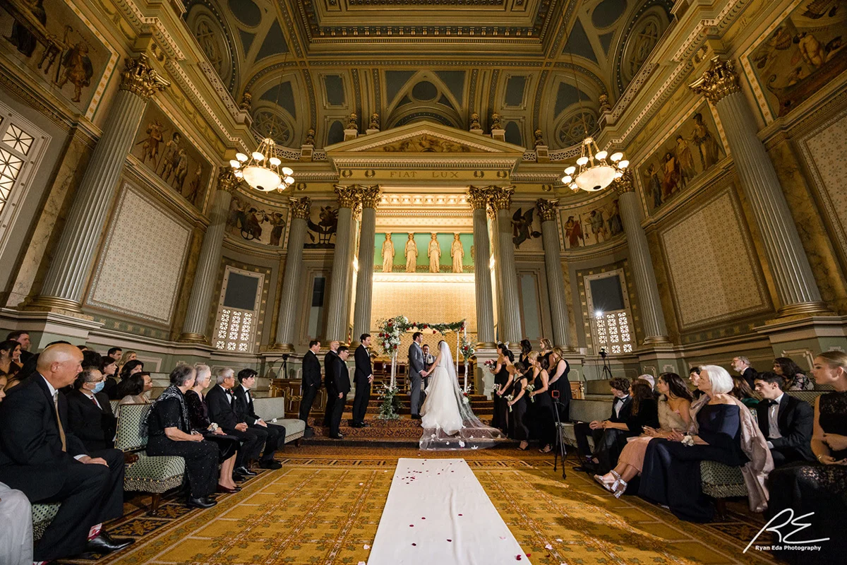 2022-10-15-Young-Wedding-427 Large Grecian-inspired room where bride and groom are standing at the front. Guests are seated on either side of the aisle.