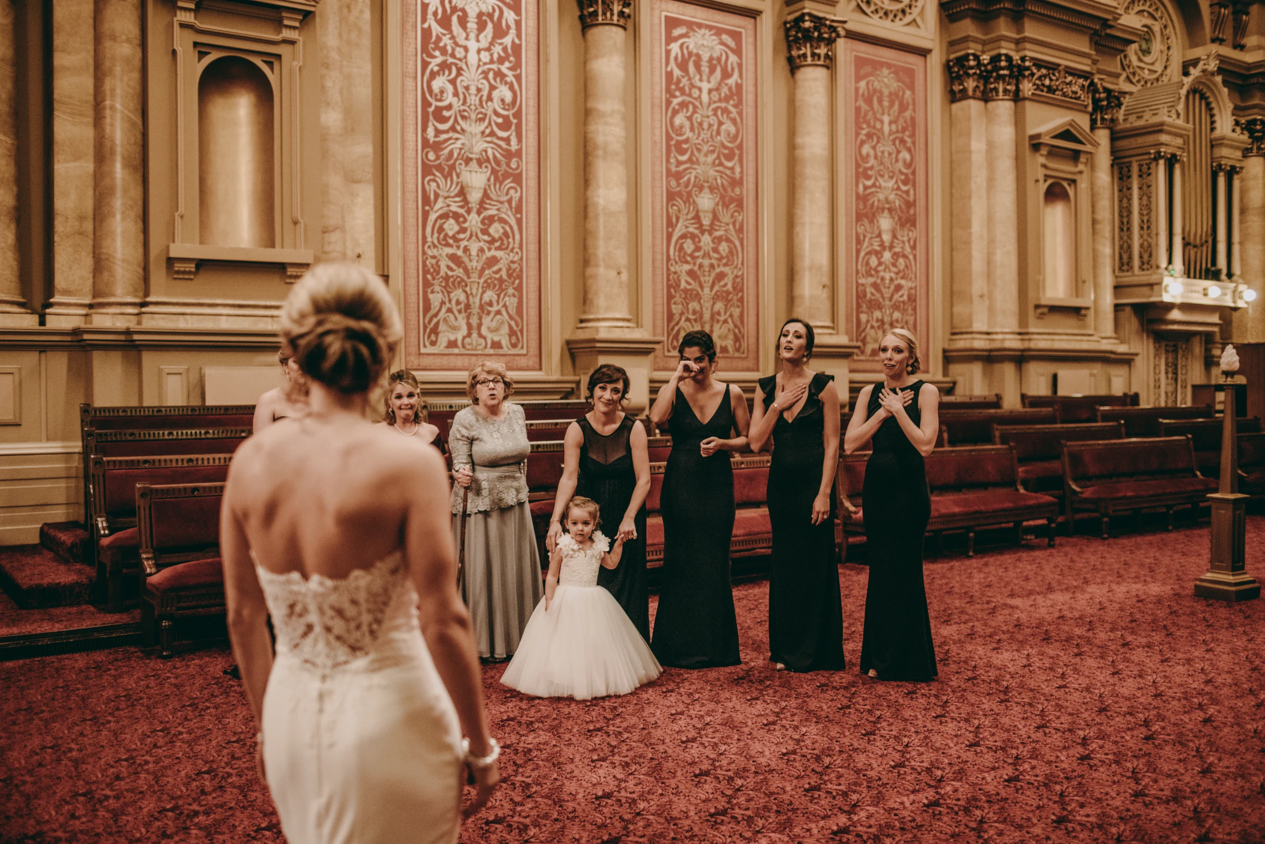 http://mariadeforrest.com Bride standing facing away from camera while her family and friends react to seeing her in a large room with cream and red accents.
