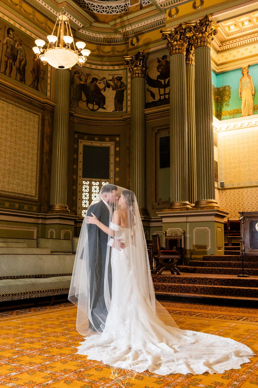 231021AM-516 Bride and groom kissing under veil while standing in large, Grecian-inspired room.