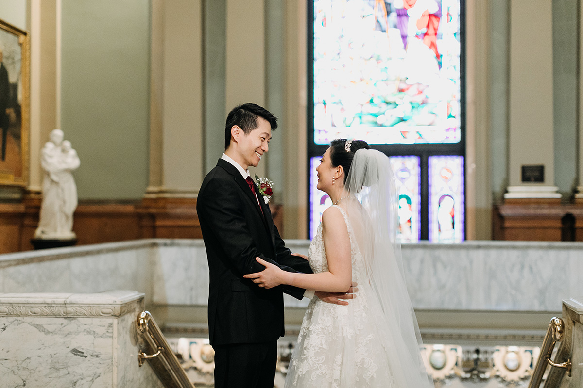 Amanda Love Me Do Smiling bride and groom holding each other with a stained glass window behind them.