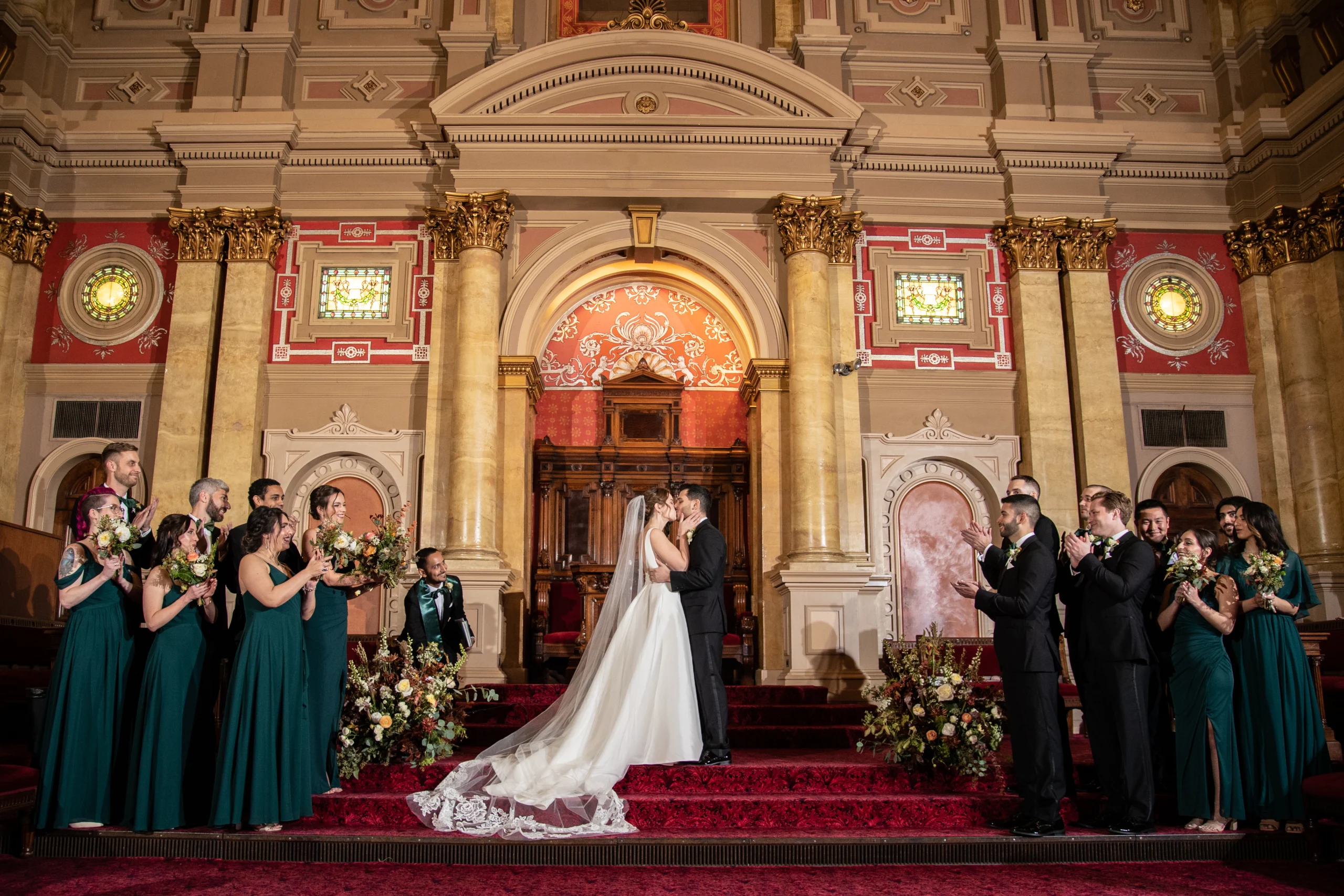 Amber Johnson Photography Kissing bride and groom standing at the front of an ornate room while bridal party applauds standing by them.