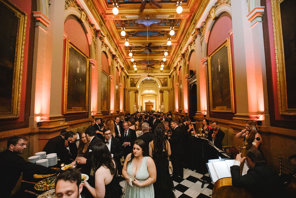 Asya Photography Large foyer with paintings on either side bustling with guests in formal attire. A food station and musicians are in the foreground.