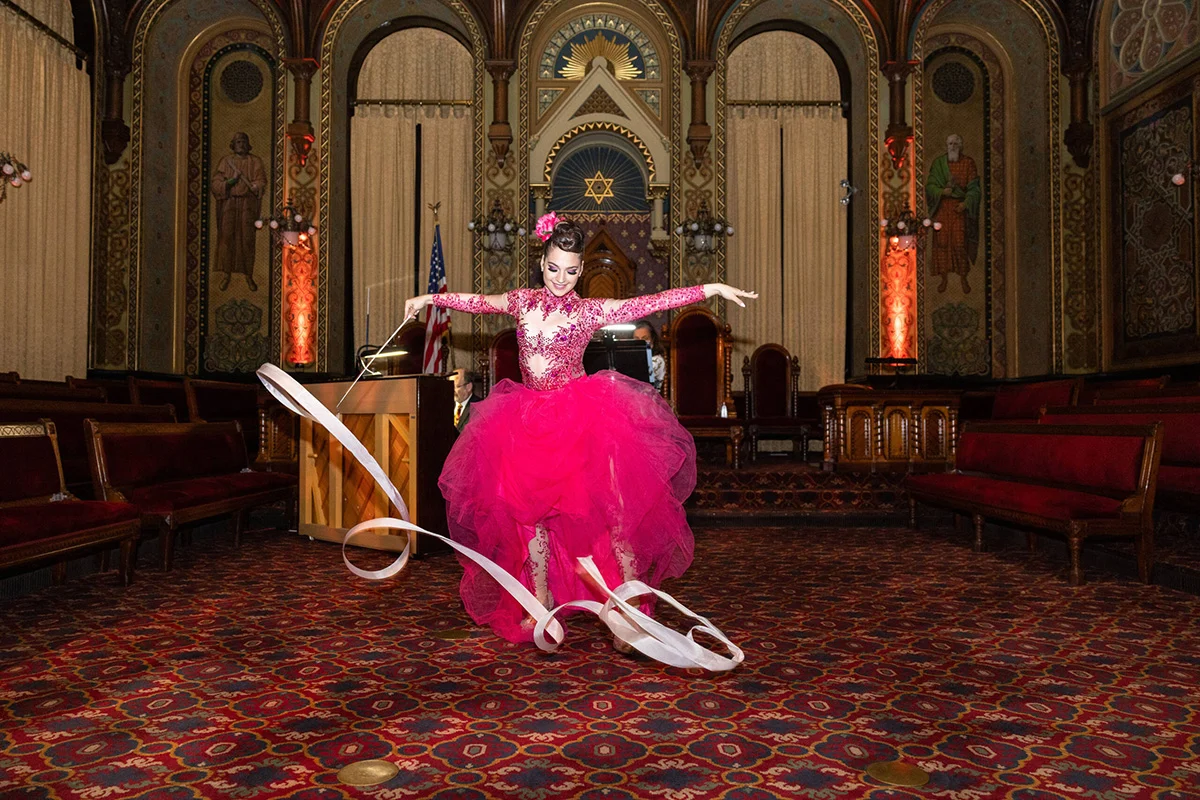 Barr_Saturday_0158-scaled Smiling woman dressed in pink dress twirling a ribbon. She's standing in an ornate room with arches and gold-accented decor.