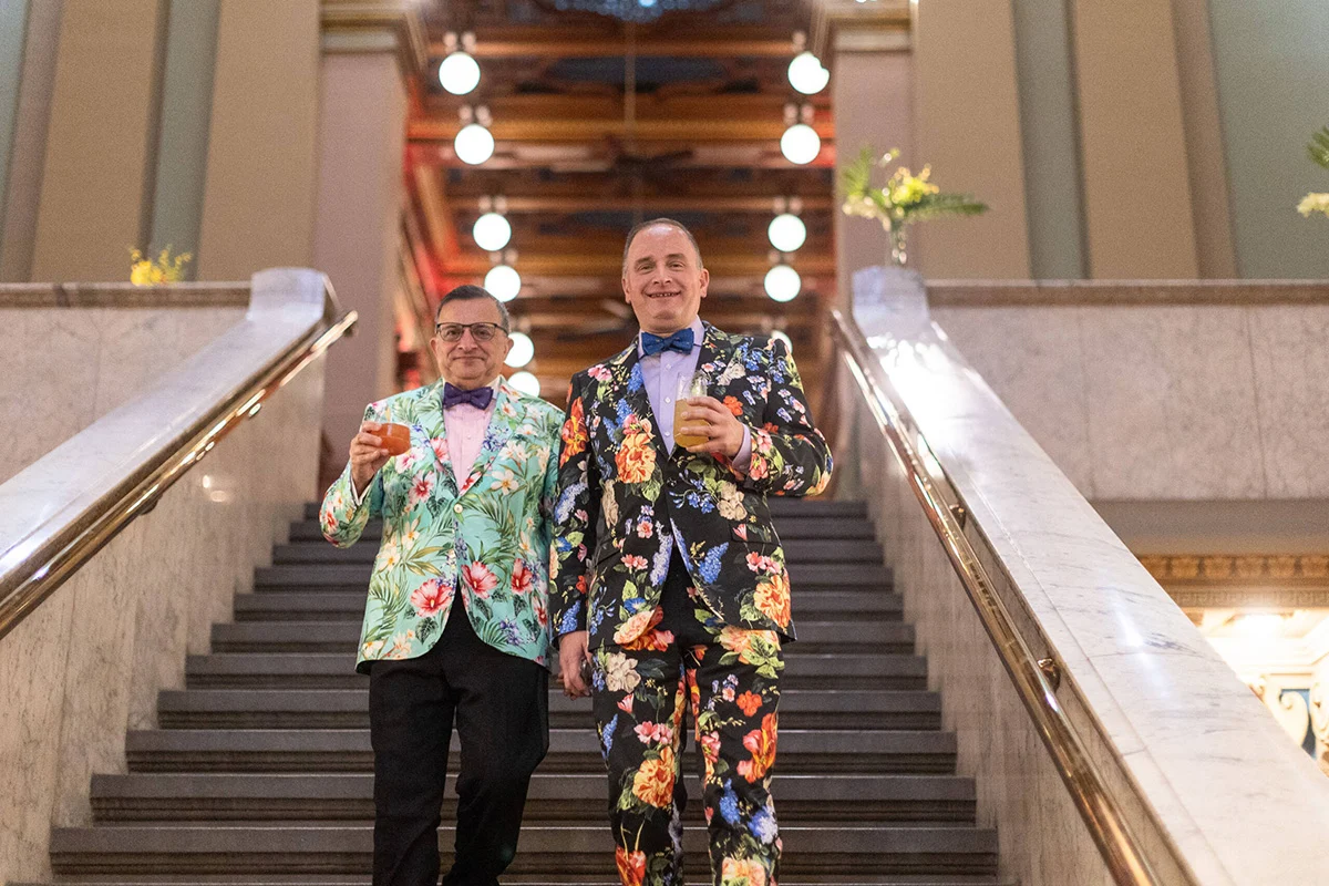 Barr_Saturday_0228-scaled Two smiling men in floral suits raising their drinks on a staircase.