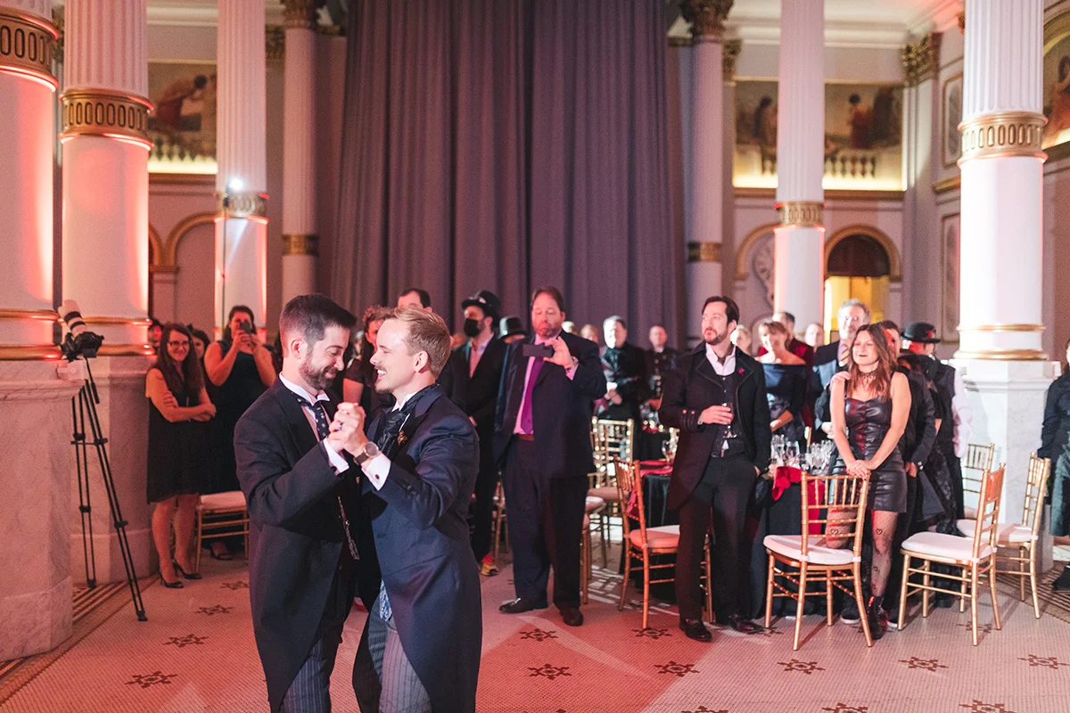 Brandon Adam Photo Smiling grooms dancing while surrounded by guests in large ballroom.