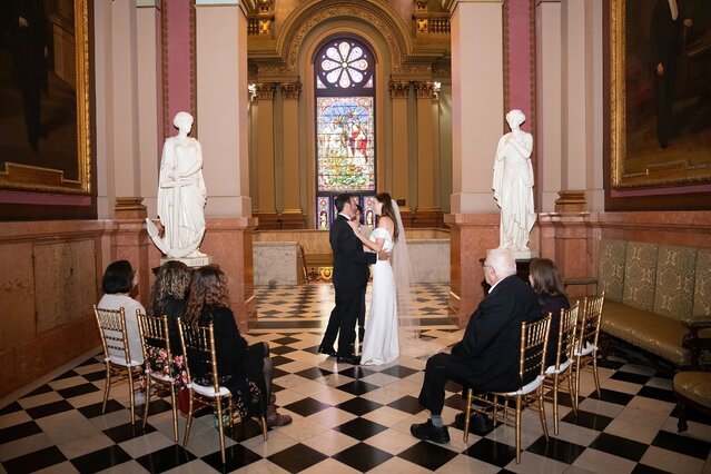 David Naples Photography (2) Bride and groom standing together in front of small crowd, with sculptures and a large, stained glass widow behind them.