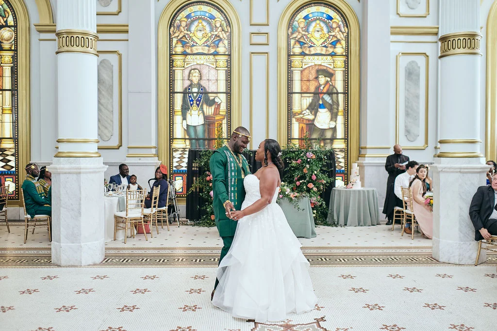 Dukk Fliks Photography (2) Smiling bride and groom dancing together in ballroom while seated guests watch. Behind them are flowers and large, stained glass windows.