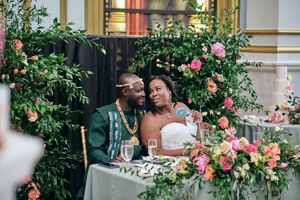 Dukk Fliks Photography Smiling bride and groom seated together at a small table while surrounded by flowers.