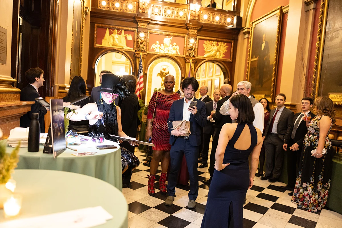 Eddie Marenco Photography (4) Group dressed in formal attire gathered in ornate foyer, there is a woman in a top hat seated to the side painting while guests are gathered near her.