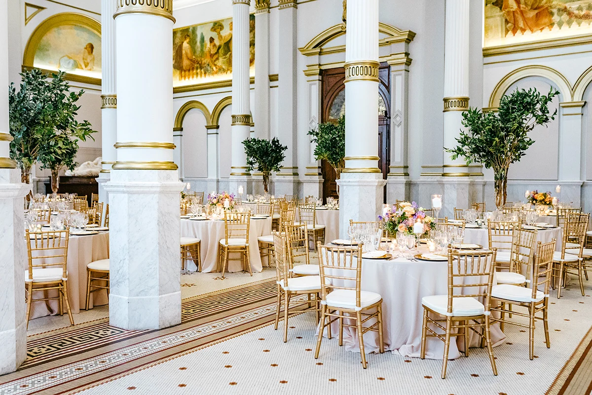 Emily Wren Photography Ballroom with round tables with white table cloths, gold and white chairs and floral displays on each table. The ballroom has large columns, tiled white and gold flooring and murals on the tops of the walls.