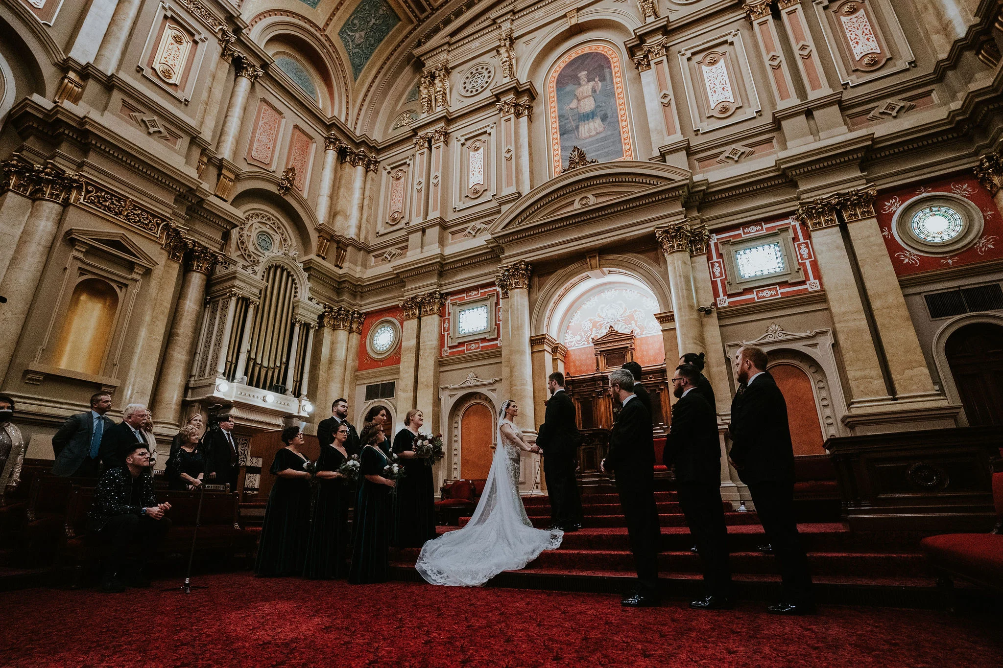 ErinSteve.OneNorthBroad.Wedding-603 Bride and groom holding hands on staircase with bridal party standing around them. A tall wall stands behind them with columns and stained glass windows.