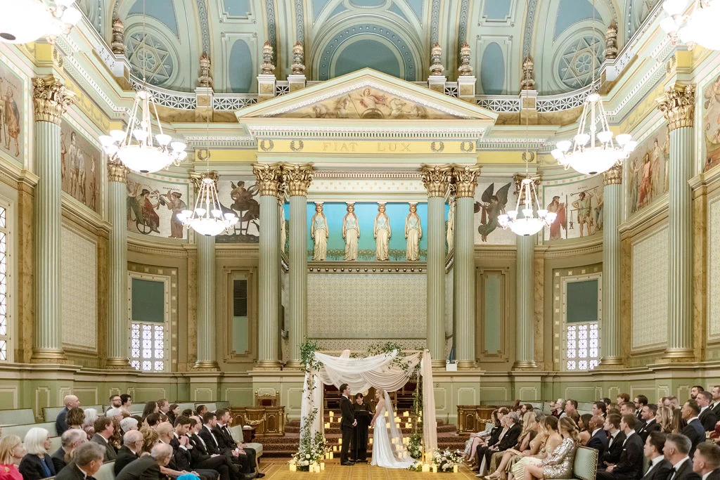 Flynn_Joe_Wedding_0848 Bride and groom being married under archway while guests sit watching from the sides. The room around them is Grecian-inspired with columns, sculptures and murals decorating the walls.