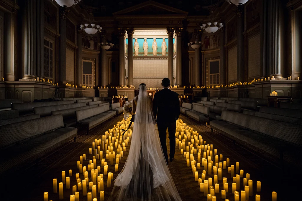 Gabrielle-Eric-211231-183359-scaled Bride and groom walking down aisle with candles on either side. In front of them, the walls are also lined with candles. Columns and sculptures decorate the walls.