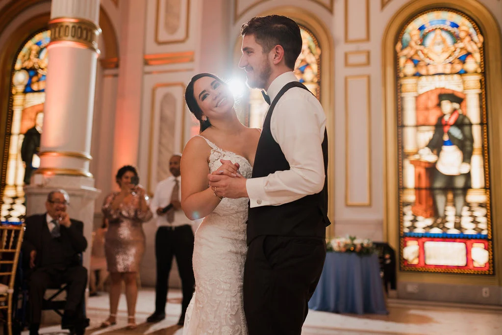 Hieu Pham Photography Smiling bride and groom dancing together in ballroom with guests, stained glass windows and a column behind them.