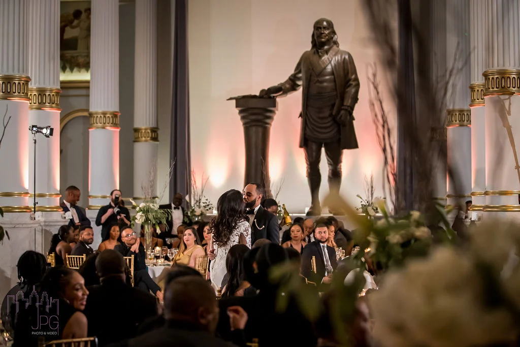JPG Photography (3) Bride and groom kissing surrounded by seated guests in a large ballroom. Columns are on either side of the room and a large statue of Benjamin Franklin is behind them.