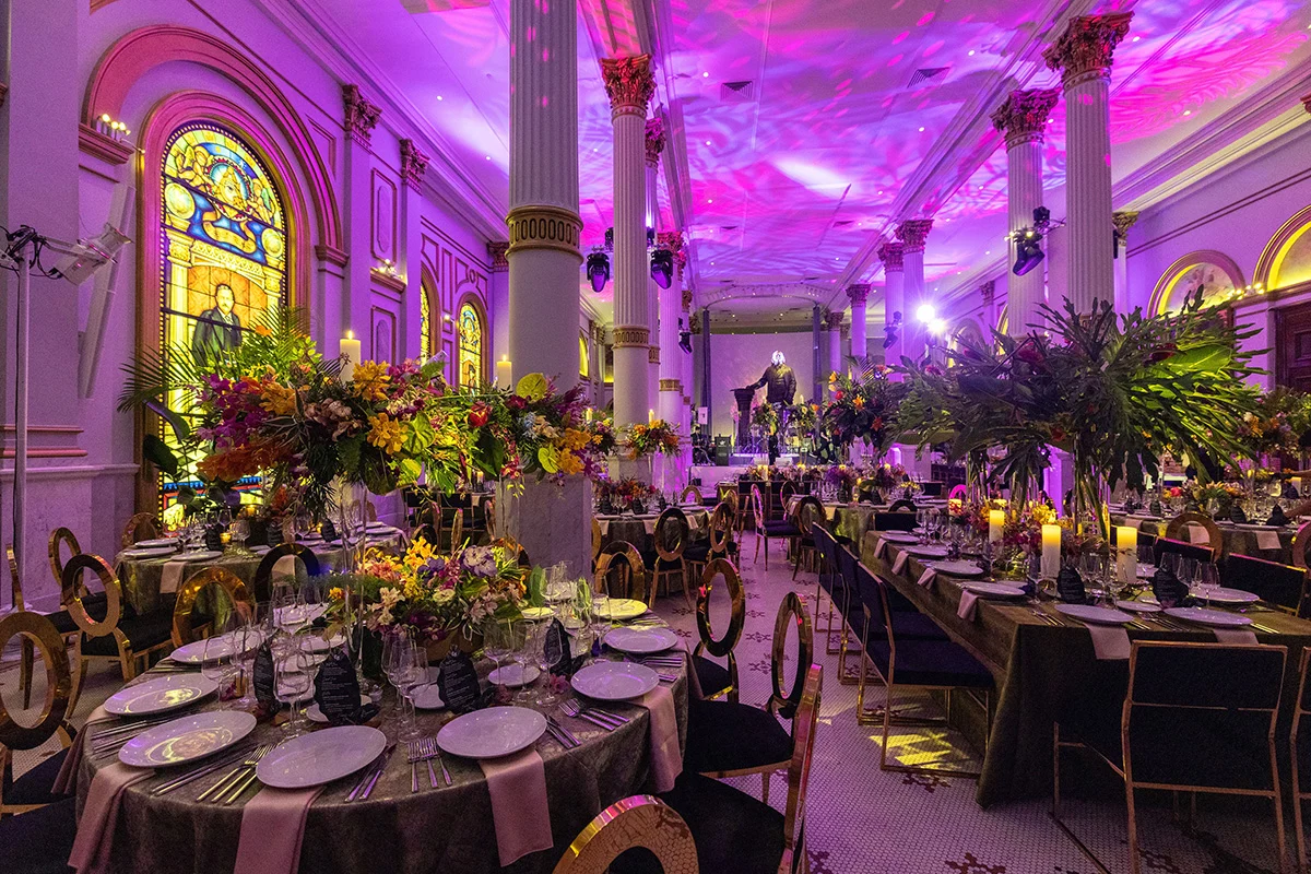 Jana Bannan Photography (5) A large ballroom filled with tables with dark table cloths, white plates and lush botanical centerpieces. Columns and a large statue are in the background and the ceiling is decorated with pink and purple swirled lighting projections.