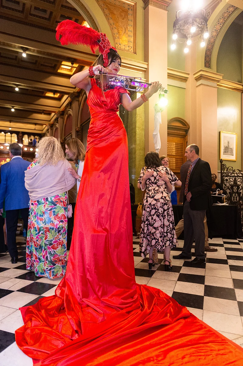 Jana Bannan Photography (6) Woman playing violin while on platform with a long, red dress extending onto the floor. A crowd dressed in party attire is gathered behind her.