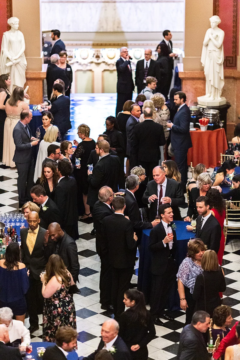 Joshua Michael Johnson Group dressed in formal attire mingling around tables with blue and orange table cloths in large foyer.