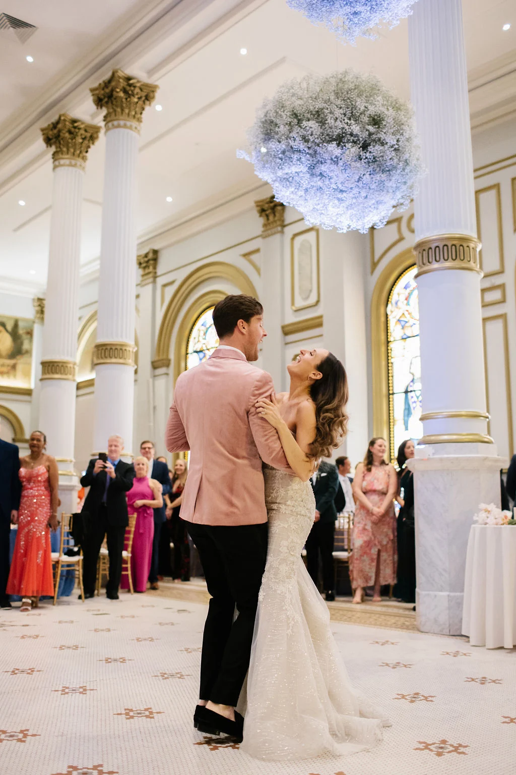 Kelly Giarocco Smiling bride and groom dancing together in Grand Ballroom. Floral displays are hanging from the ceiling behind them and guests are gathered in the background.
