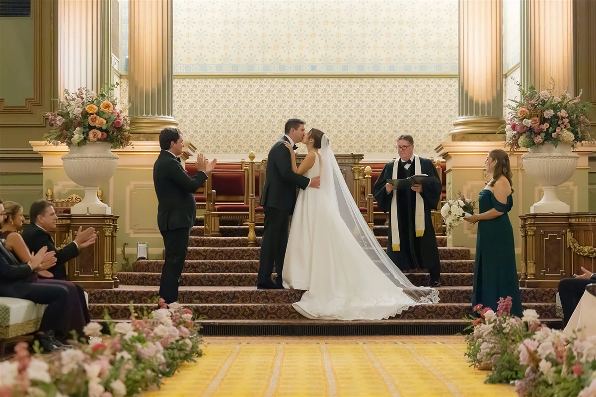 LM-610 Bride and groom kissing on staircase while officiant and wedding party stand to the side. Flowers are on either side of them and the aisle is lined with matching flowers.
