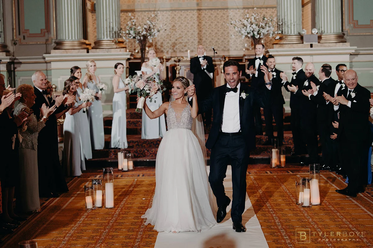 OneNorthBroad.KristinChris.wedding.tylerboye-65-scaled Smiling bride and groom walking together as wedding party and guests celebrate around them.
