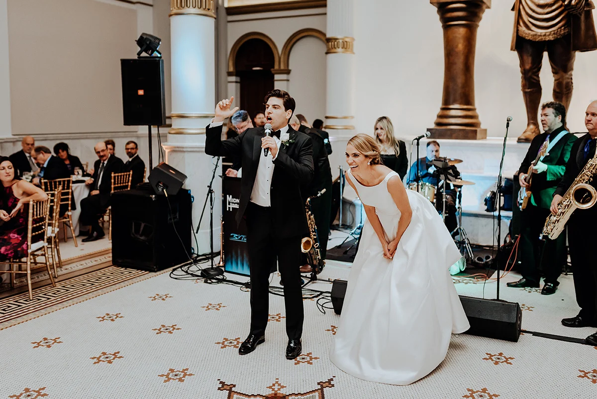 Pat Robinson Man in suit and bowtie speaking into microphone while laughing bride is standing beside him. Guests are watching from tables while band stands behind them.