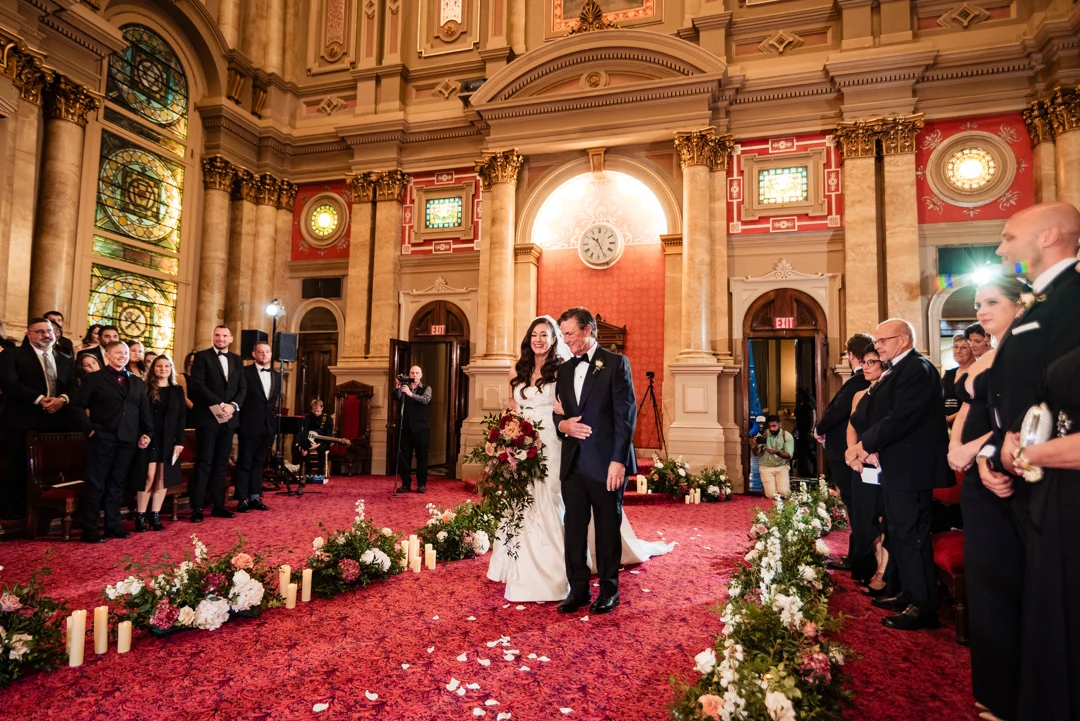 Ralph Deal Photography8 Bride and her father walking down aisle together lined by candles and flowers while guests watch on either side. The room is large with cream walls, stained glass windows and red carpet.