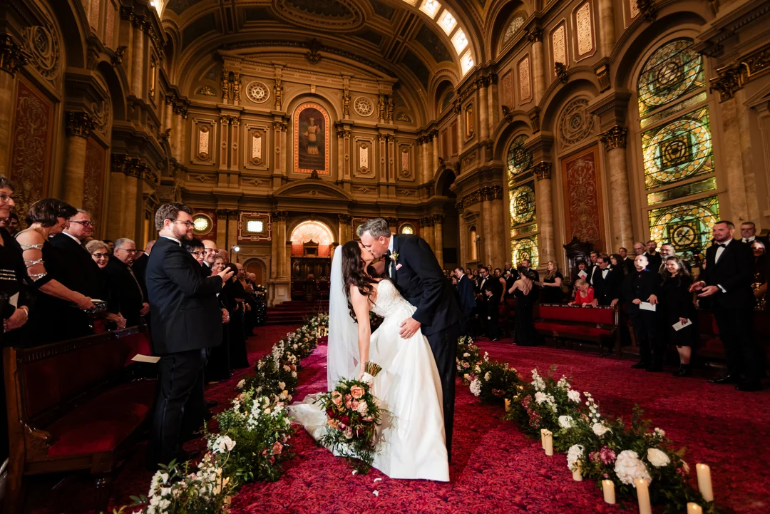 Ralph Deal Photography9 (2) Bride and groom kissing in center of flower-lined aisle while guests applaud behind them. The room around them has tall ceilings, stained glass windows and ornate, cream colored walls.