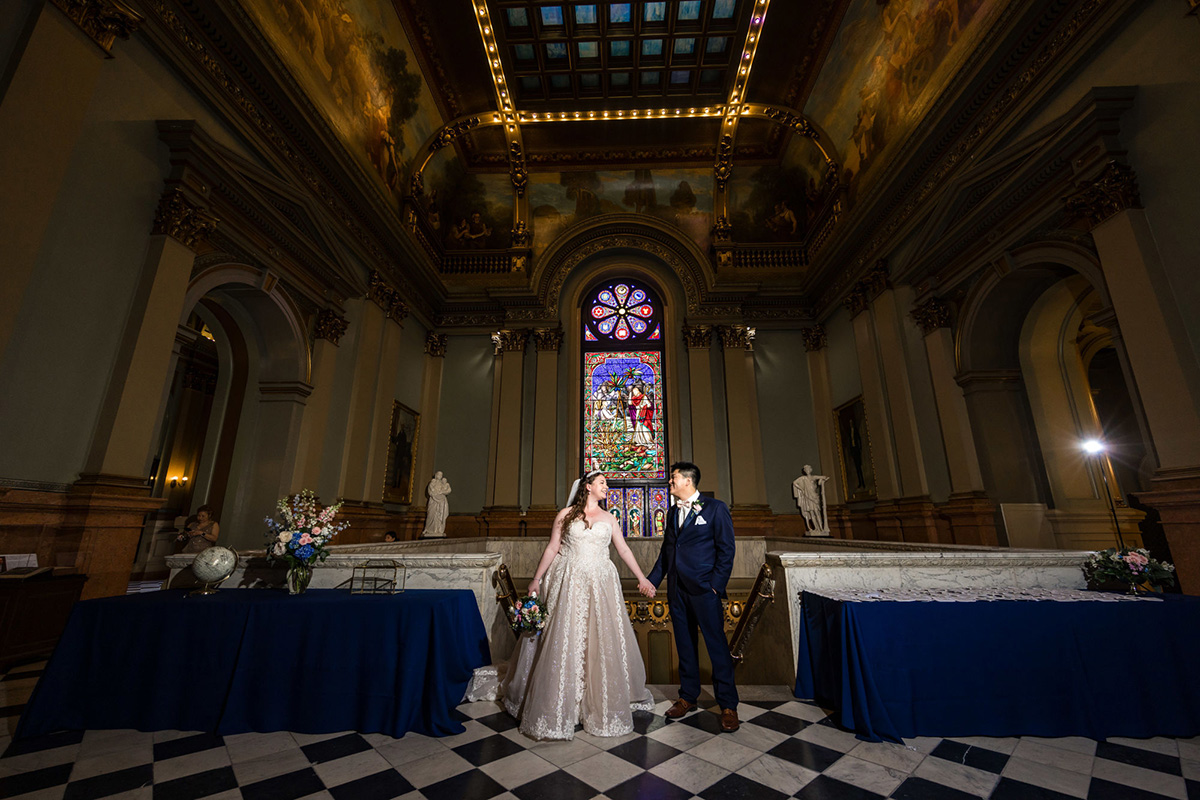 Ryan Eda Photography (2) Bride and groom holding hands in large foyer with ornate walls and stained glass windows behind them. Tables with blue cloths are on either side of them.