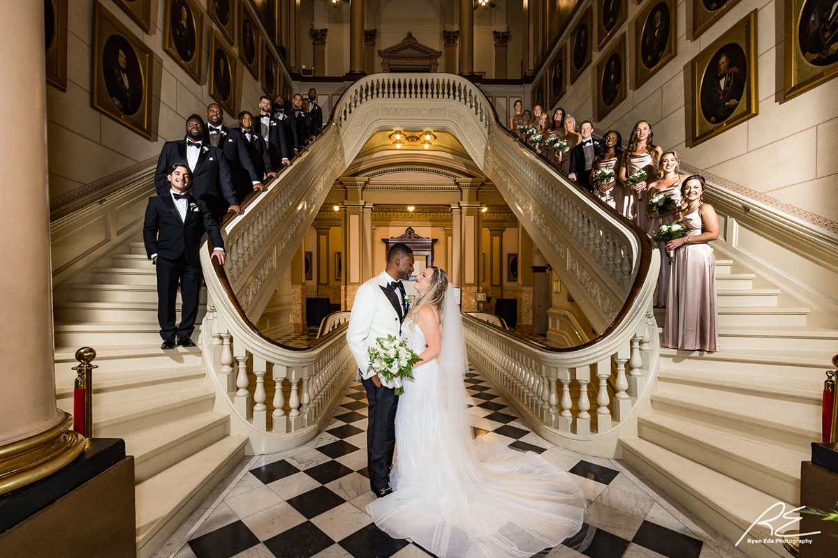 Ryan Eda Photography (4) Bride and groom standing together in front of double staircase while smiling bridal party stands behind them on the staircase.