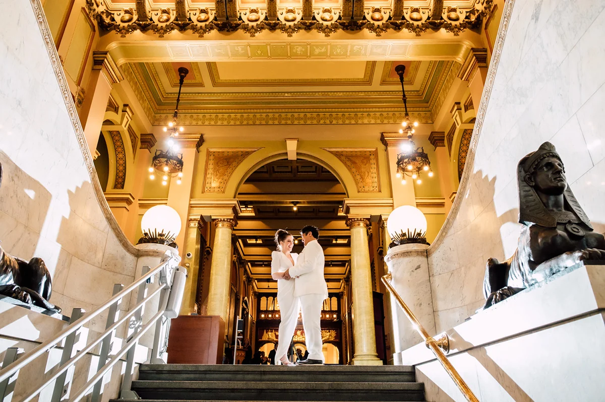 Sean Marshall Lin Smiling couple dressed in white at the top of an ornate marble stairway.