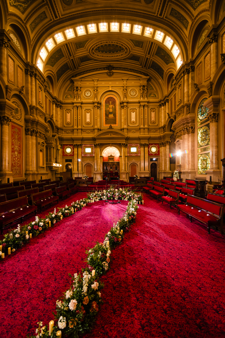 Hall with high ceiling and ornate walls with flowers laid out in an aisle down the center.