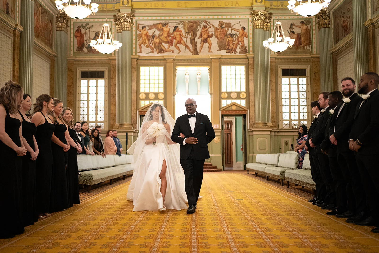 Matt & Liana Smiling bride walking down aisle with her father while guests and bridal party stand on either side. The room around them is large and Grecian-inspired with high ceilings, chandeliers and a gold-colored carpet.