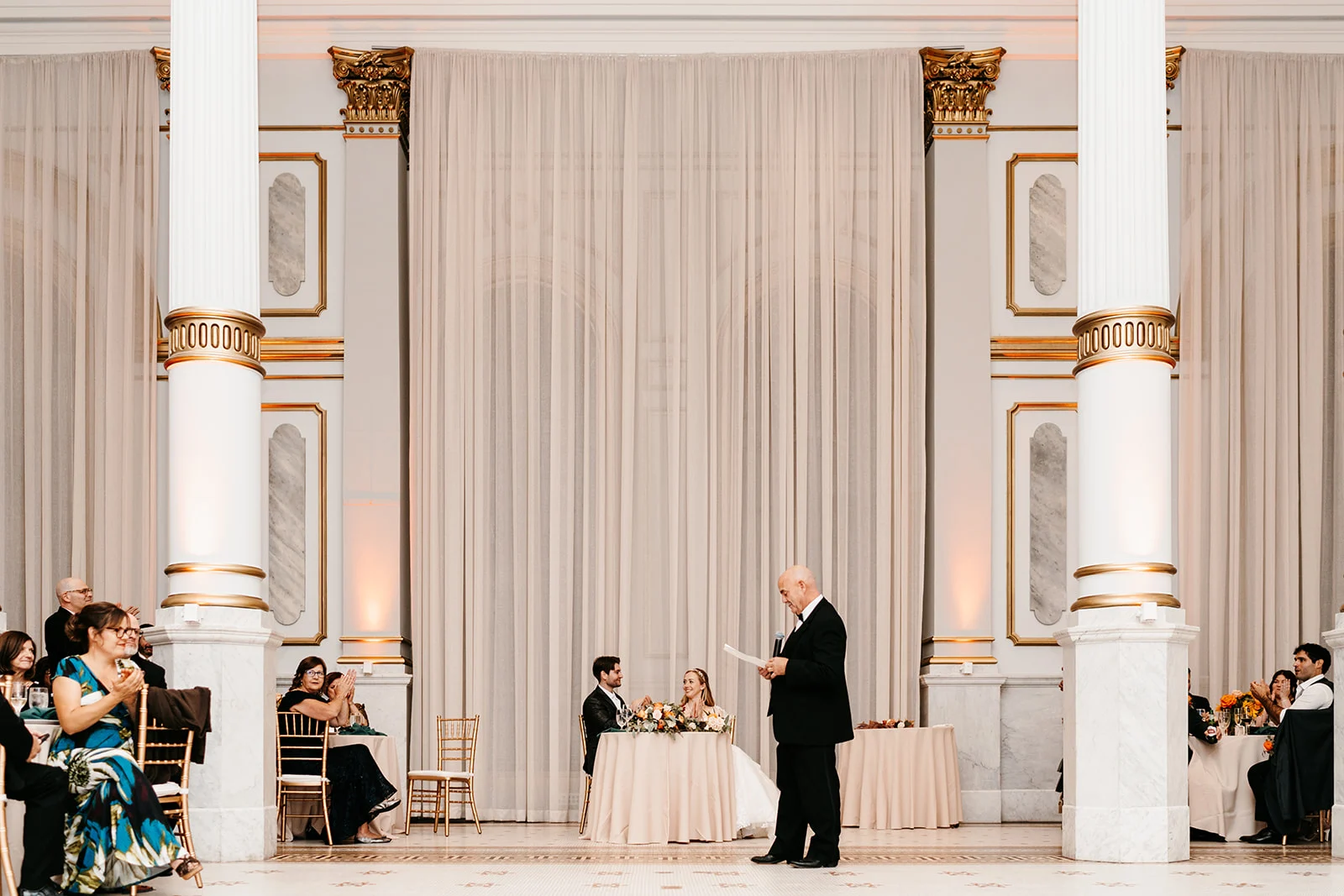 Nick and Sarah Man giving speech in large ballroom while bride, groom, and wedding guests applaud from their tables.
