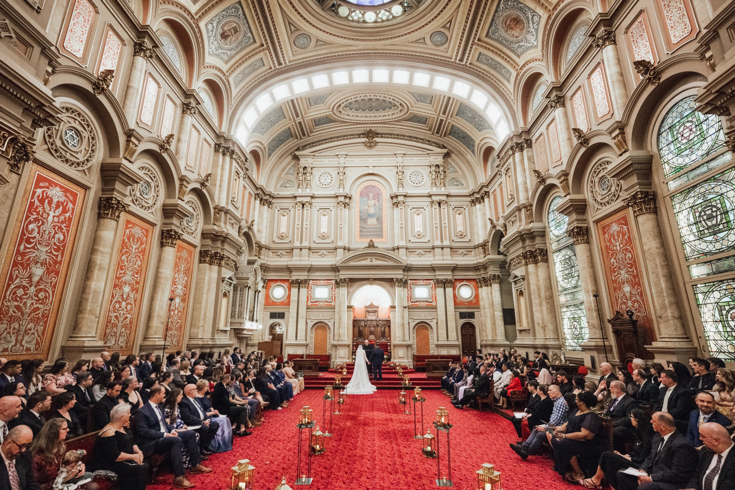 Ted Felsberg Photography3 A large room hosting a wedding. The room has tall ceilings and stained glass windows. A bride and groom stand together at the front of the room, while guests watch from either side of the room.