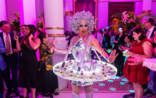 Smiling woman in silver outfit with small appetizers set on a suspended tray surrounding her.