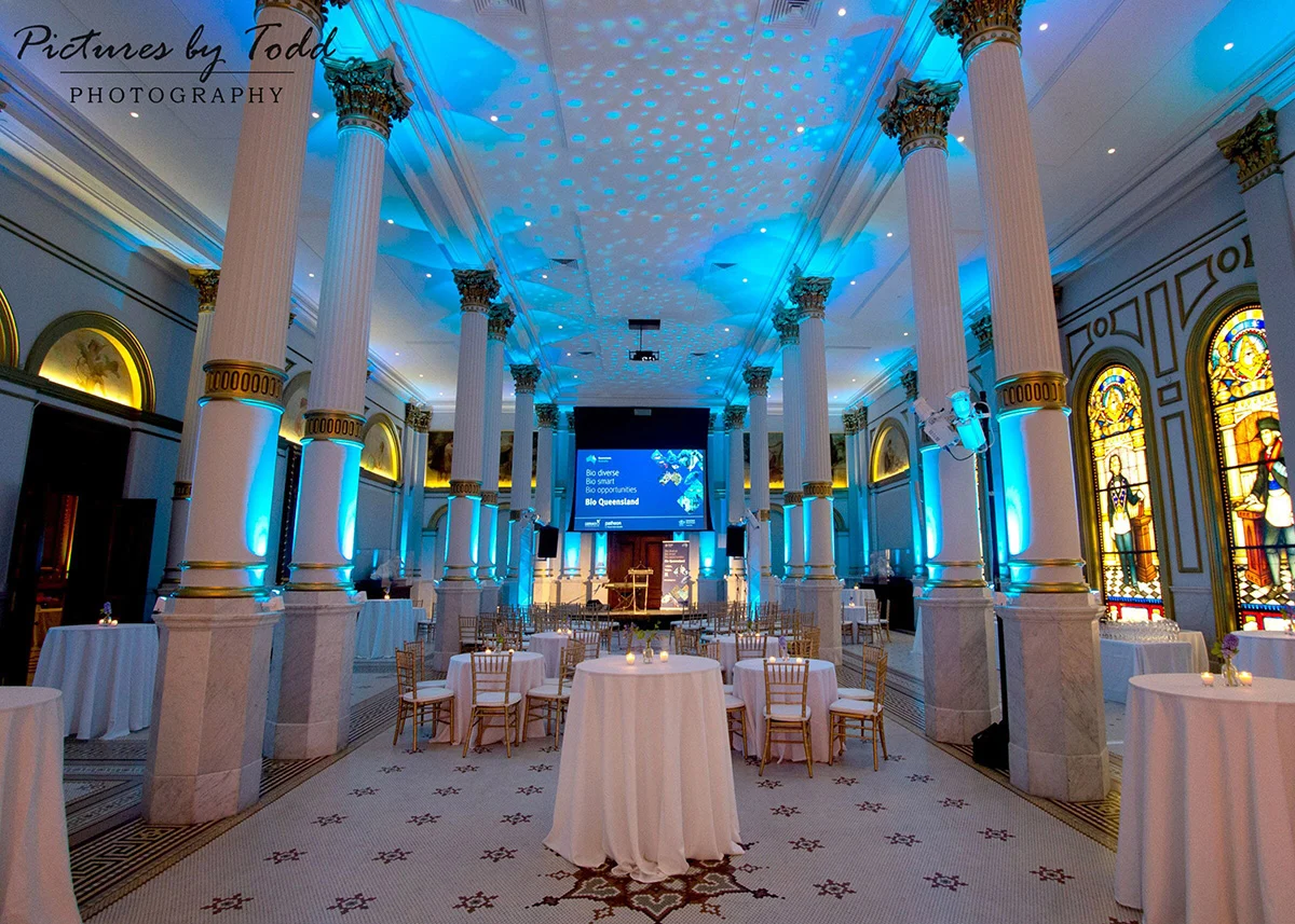 pictures by todd Large ballroom with small circular tables and gold and white chairs set for guests. The room is lit with light blue accents and pattern on the ceiling.