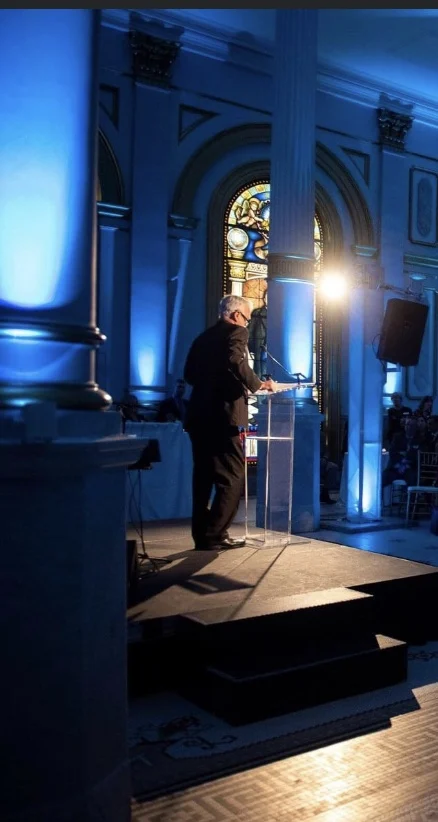 Screenshot Man in suit speaking at podium surrounded by columns illuminated in blue.