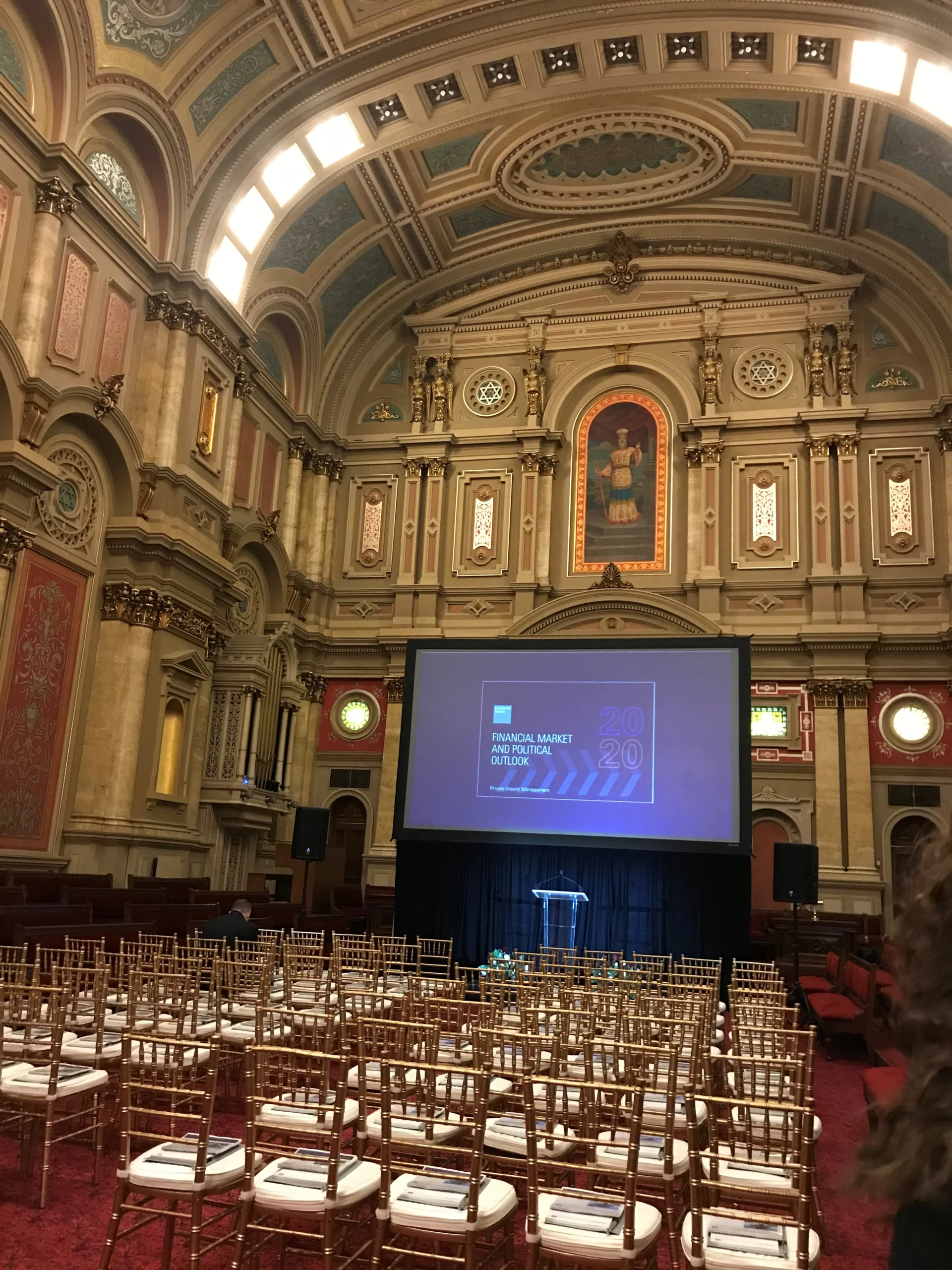 staff photo Large screen set up with visual display on a black stage with a glass podium in front of it. Empty chairs are set up in rows facing the stage. The room is large with a tall ceiling, ornate stained glass windows and a mural featuring a man at the front.