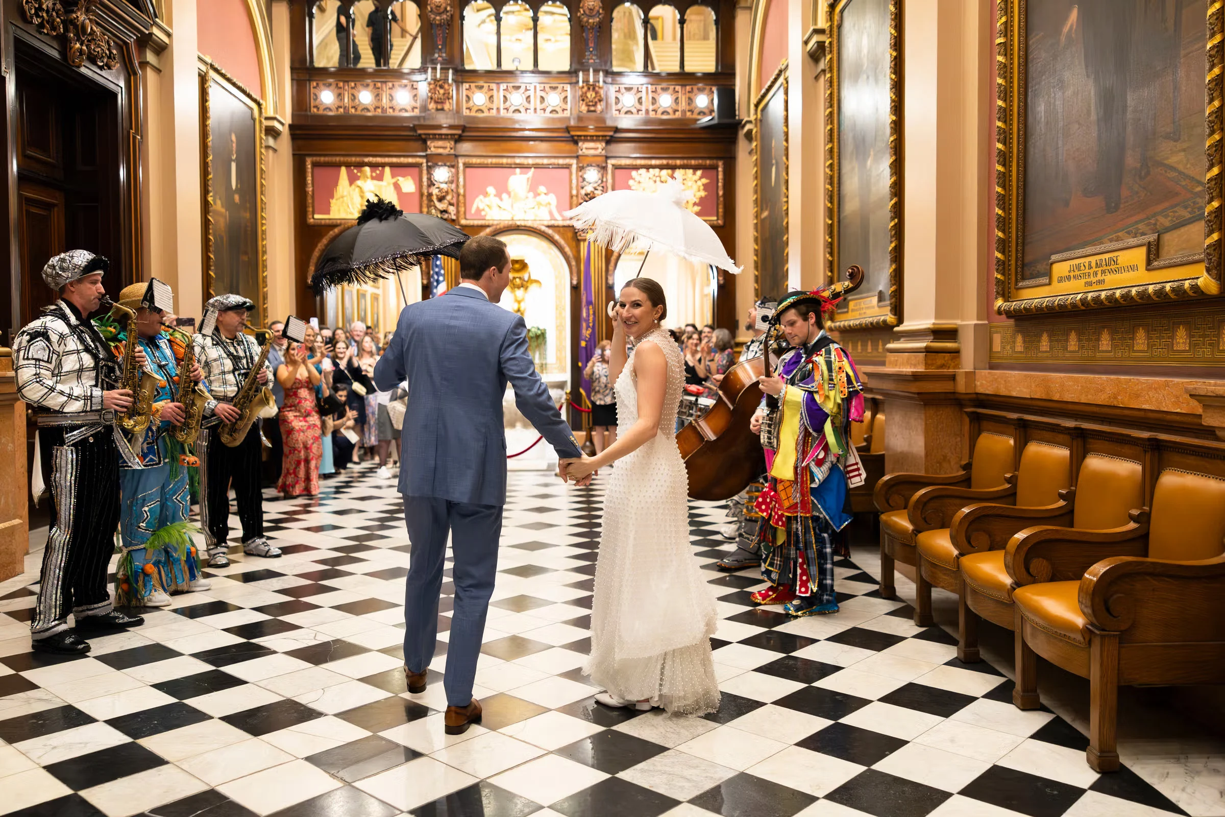 5d0afe01-0290-4652-941a-c5c6dff2db22-ps (1) Bride and groom holding feathered umbrellas while holding hands. A Mardi Gras band is playing music on either side of the foyer while they walk down the center.