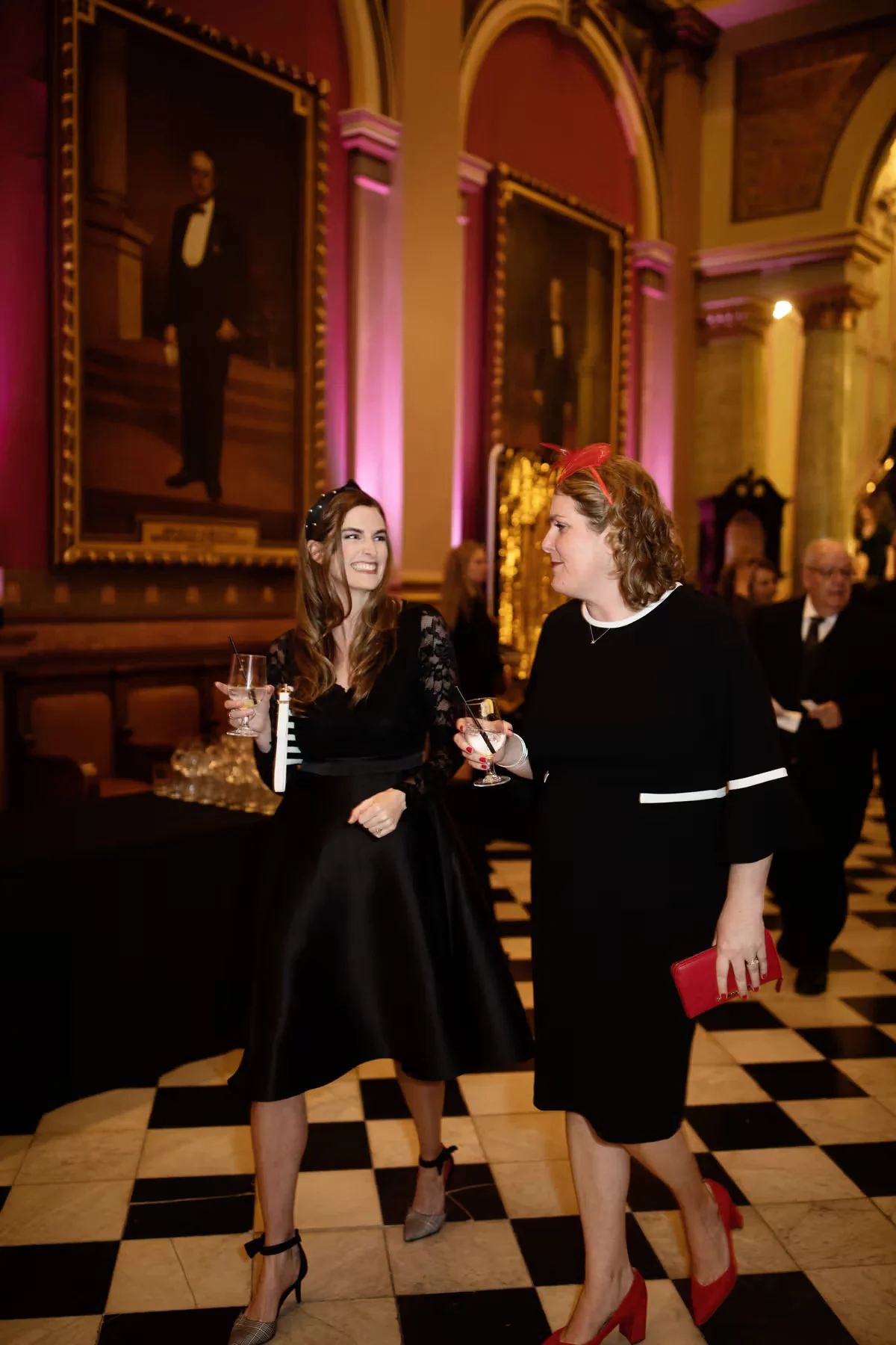 photo-d46ab3ea-b102-4047-a2cd-9da001669a24 (1) Two smiling women, dressed in formal business attire, walking together while holding glasses of wine. Paintings of men on walls with arches are behind them.